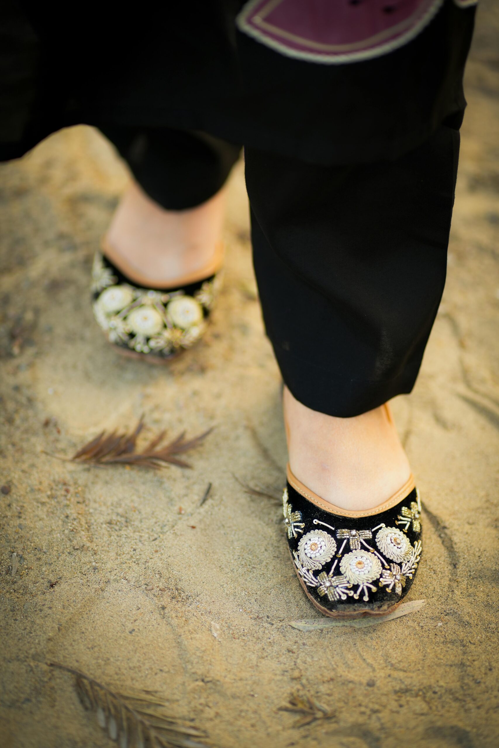 Close-up view of embroidered black shoes worn on a sandy outdoor surface, showcasing intricate floral patterns.
