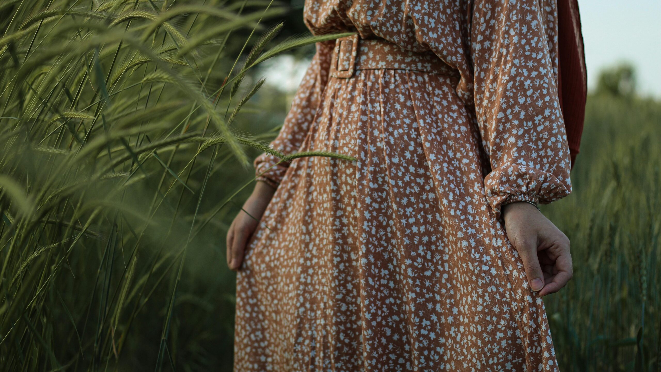 Unrecognizable crop female in elegant dress standing in grassy meadow near tall lush green grass in countryside