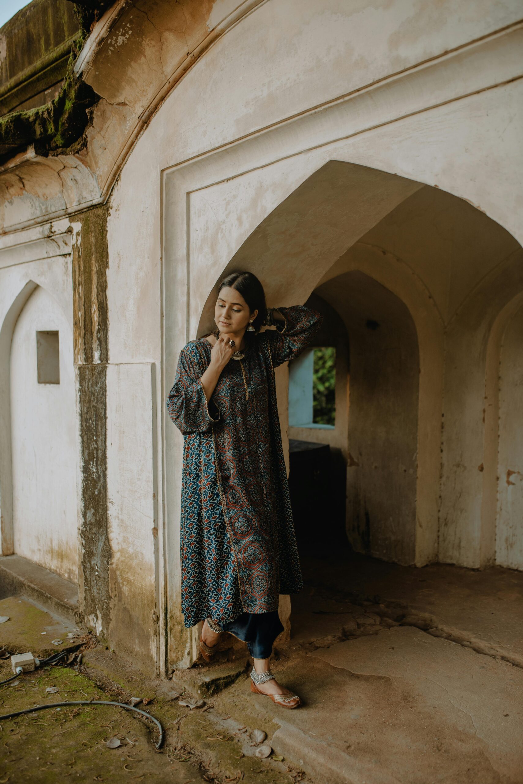 Full body of young female in sandals and traditional dress and trousers of Muslims clothes of Central Asia leaning on stone wall in arched vaults